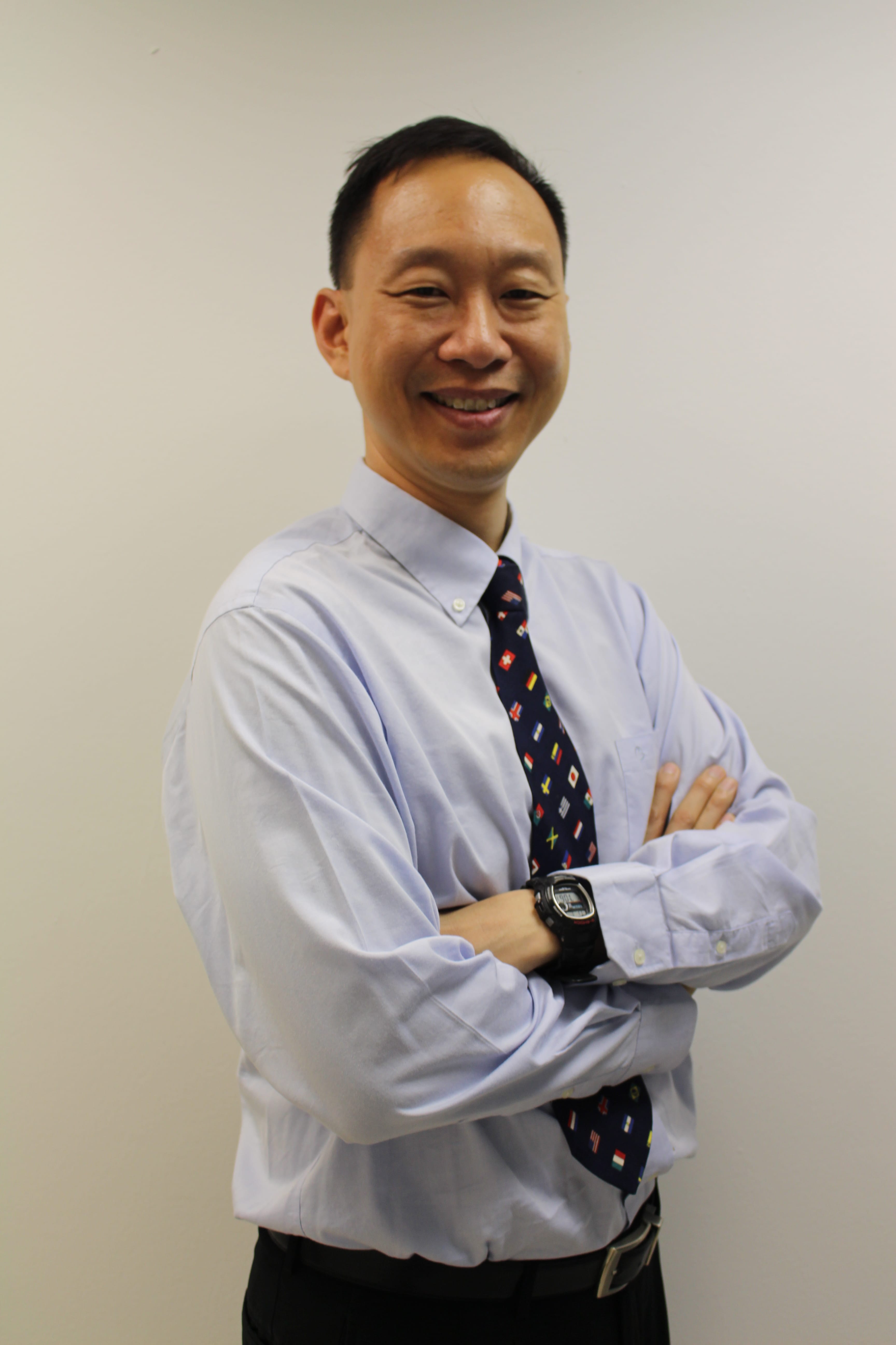 A man stands with his arms crossed, smiling warmly at the camera. He is wearing a light blue long-sleeved shirt and a navy blue tie patterned with small national flags. He has short dark hair and is wearing a black digital watch. The background is a plain light grey wall.